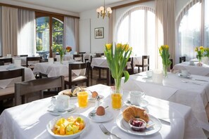 Un comedor con mesas preparadas para el desayuno, mostrando fruta, croissants y zumo de naranja.