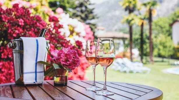 A table with a bottle of wine and two glasses, set against a backdrop of vibrant flowers and a mountainous landscape. - Garni Elena (Losone)