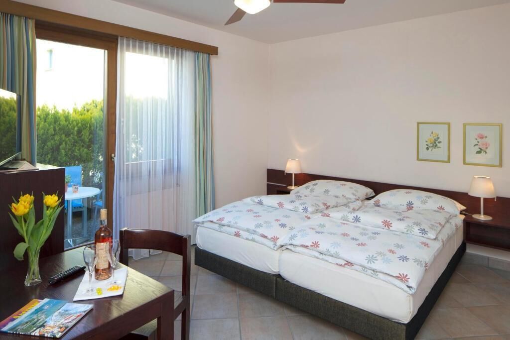 Habitación de hotel con cama, escritorio con botella y vasos, una silla y vista a zonas verdes por la ventana.