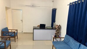 Ein Zimmer mit einem blauen Sofa, zwei Holzstühlen, einem kleinen Tisch mit einem Laptop, einem dekorativen Gegenstand und einem Vorhang.