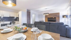Een moderne eetruimte met een houten tafel gedekt voor een maaltijd, een grijze bank op de achtergrond en een keuken met witte kasten en een zwart aanrecht.