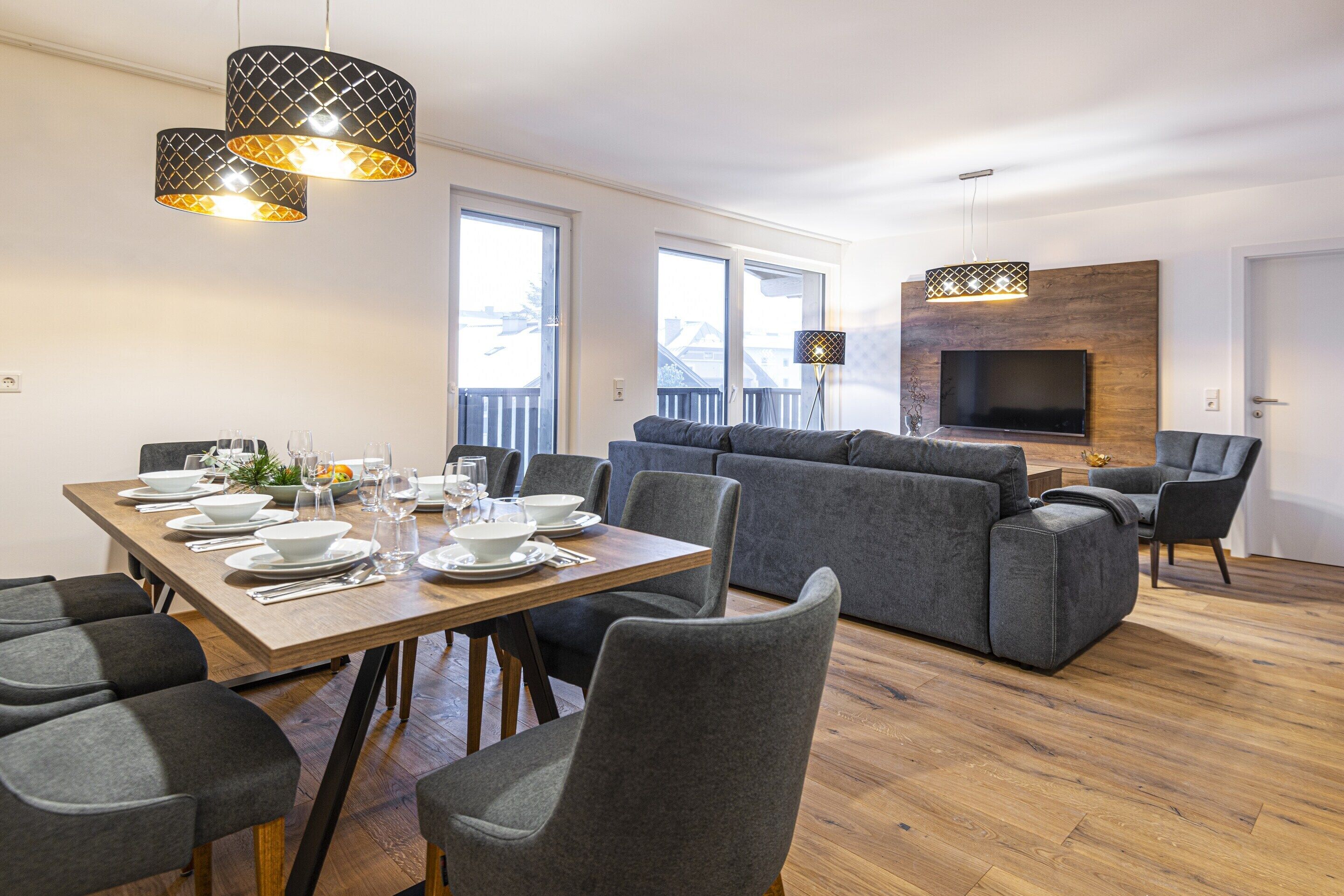 A modern living room with a grey sofa set, a wooden dining table set for four, and a flat-screen TV mounted on a wooden wall.