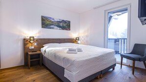 Een slaapkamer met een groot bed, een houten hoofdeinde, bedlampjes, een stoel en een raam met uitzicht op een berglandschap.
