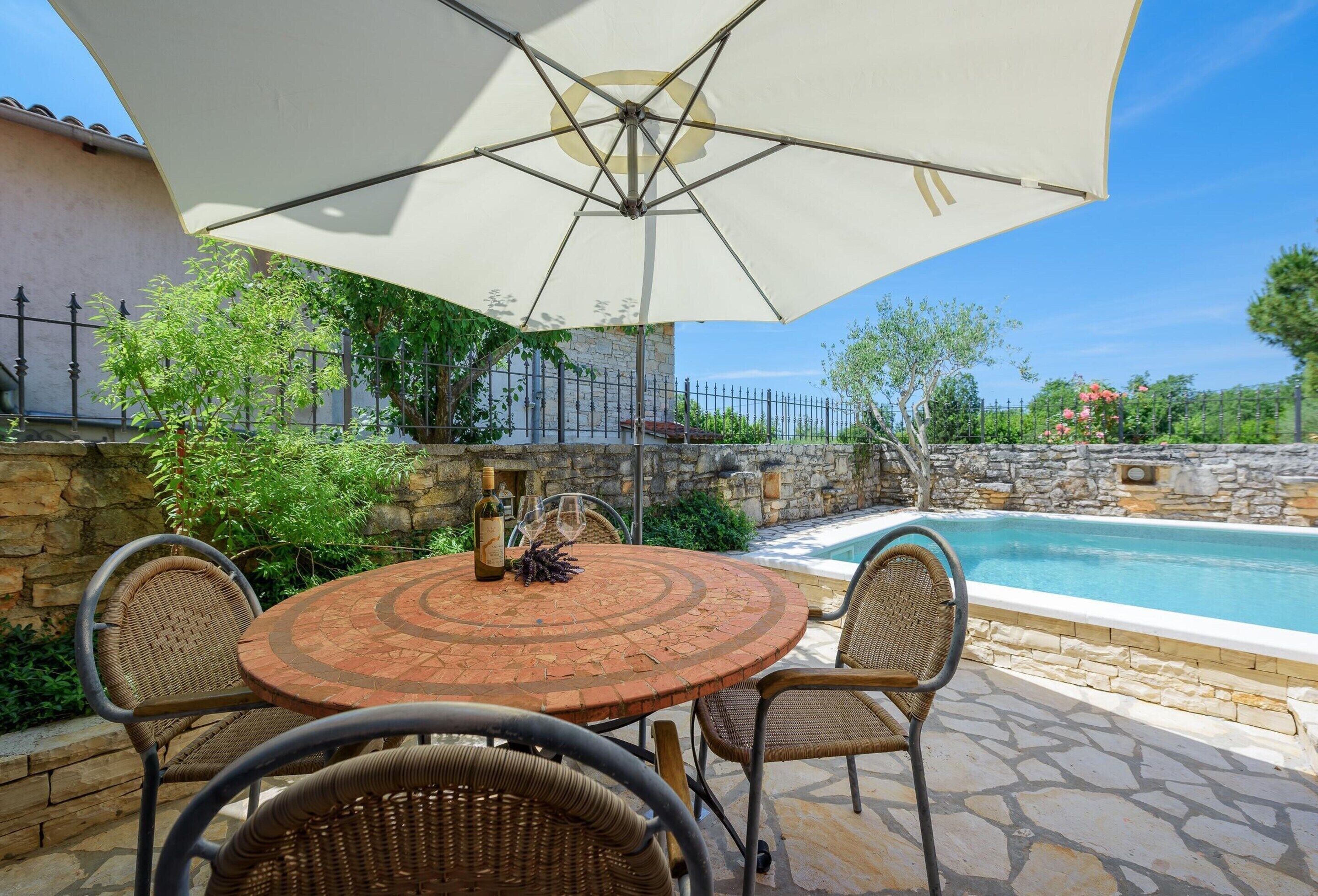 A round table with wicker chairs and a large umbrella, set against a stone wall and a pool.