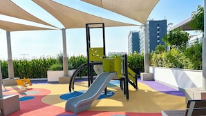 A children's play area with a slide and climbing structure, surrounded by a covered seating area and colorful flooring. - Urban Charm: Studio With Balcony in Riviera (Dubai)