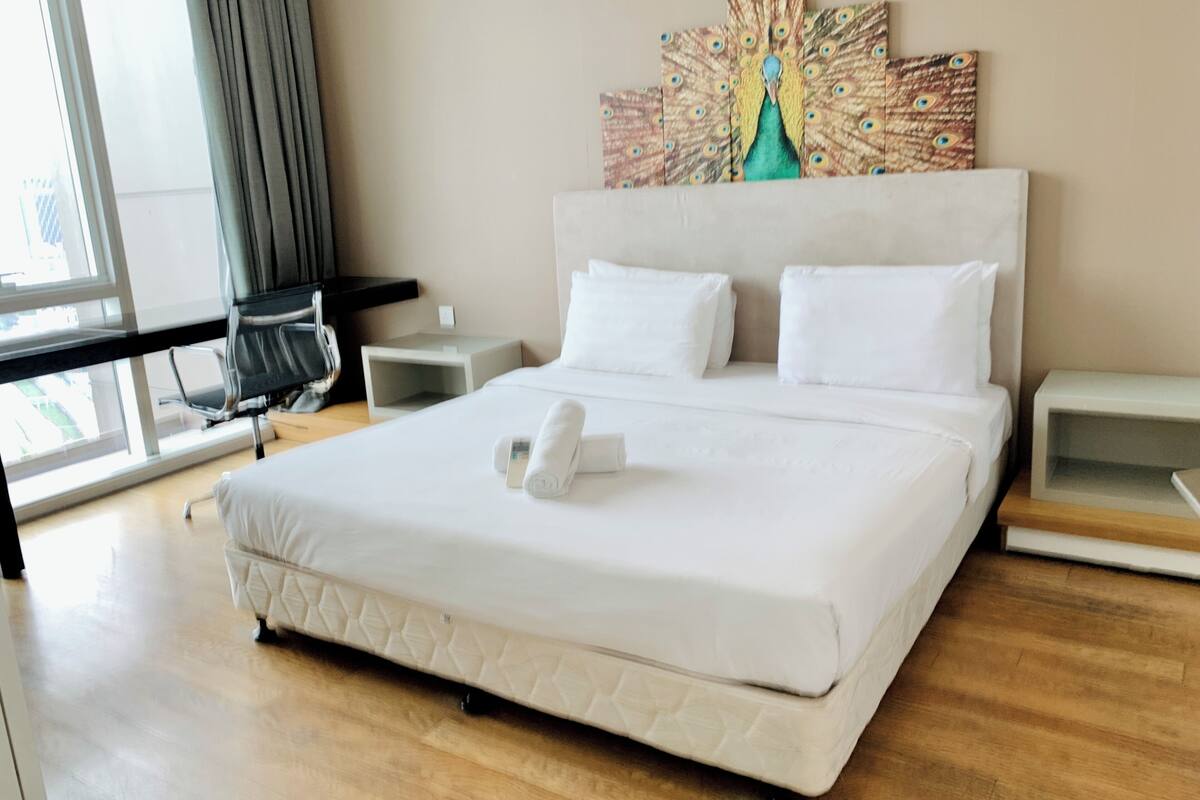 Une chambre d’hôtel avec un grand lit, deux tables de chevet, un bureau avec une chaise, un tableau représentant un paon accroché au mur et une fenêtre avec des rideaux.