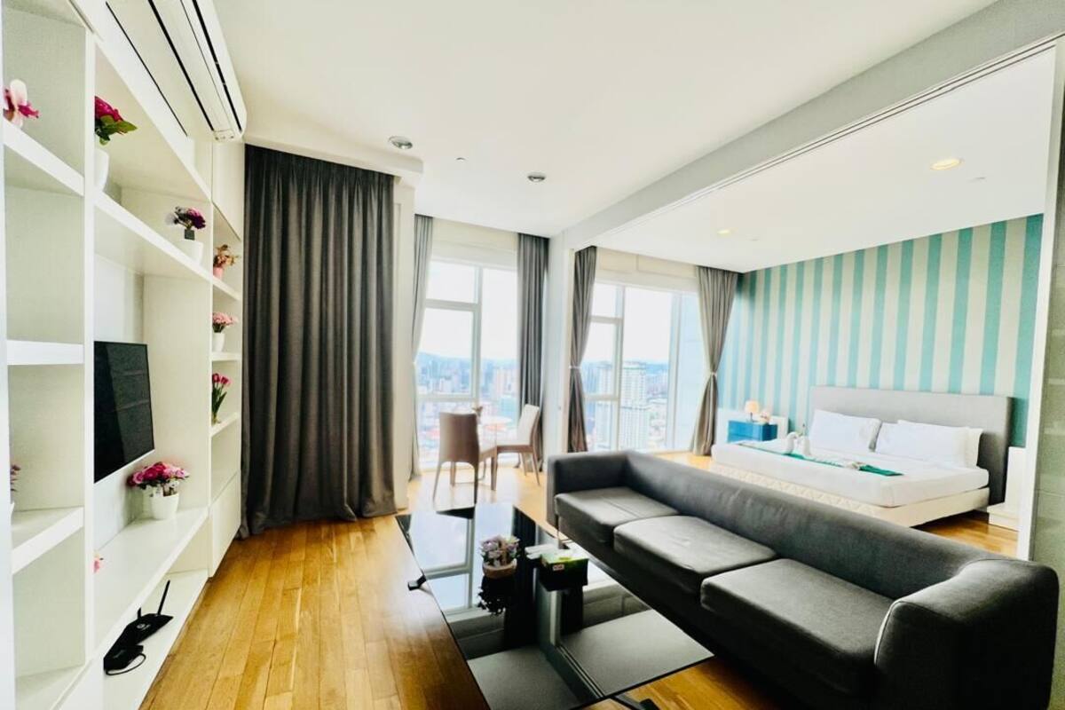 Une chambre d’hôtel moderne avec un canapé gris, un lit avec du linge de lit blanc, un téléviseur posé sur une étagère blanche, un bureau avec une chaise et de grandes fenêtres avec des rideaux.