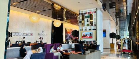 A modern hotel lobby with seating areas, a reception desk, and a television screen.