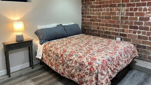 A bedroom with a brick wall, a bed with floral bedding, a bedside lamp, and a small wooden nightstand. - Private Garden Unit Gary BP (Gary)