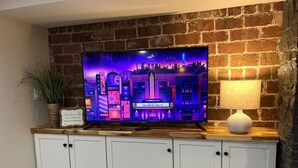 A television on a brick wall, a small table with a lamp, and a cabinet below. - Private Garden Unit Gary BP (Gary)