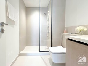 Et moderne badeværelse med et glasafskærmet brusebad, et hvidt toilet og et håndvaskarrangement med et spejl og en vase med blomster.