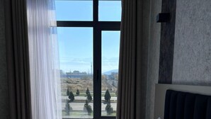 A hotel room with a large bed, a view of a landscape with trees and buildings, and sheer curtains on the window. - K HOTEL RESTURANT (Bishkek)