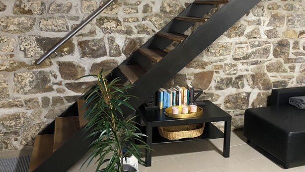 Uma escadaria com parede de pedra ao fundo, um vaso de planta e uma pequena mesa com livros e velas.