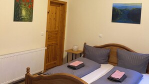 Quarto com cama de madeira, duas pinturas nas paredes, porta de madeira e uma pequena mesa de cabeceira com abajur.