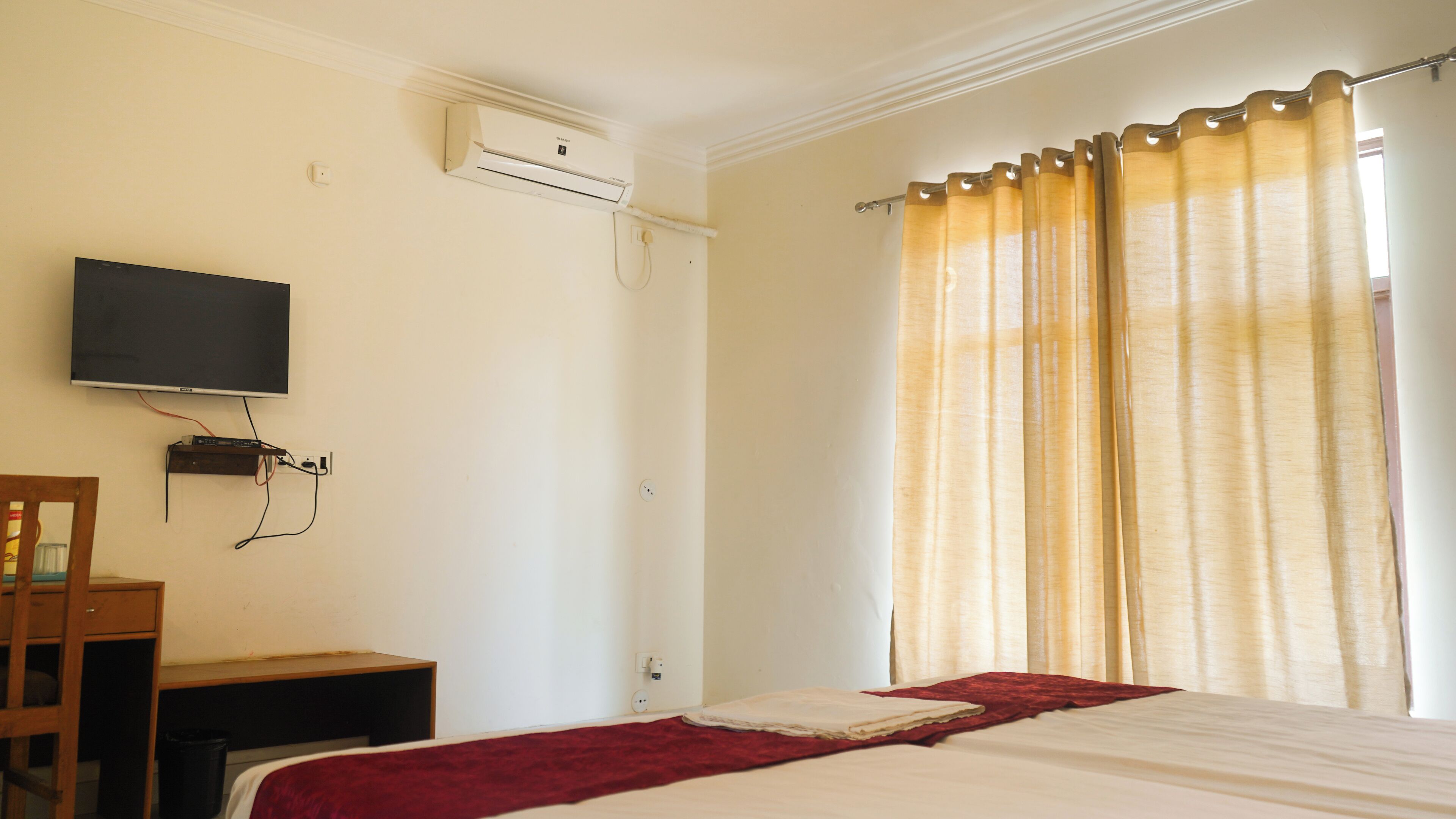 A bedroom with a bed, a TV mounted on the wall, a desk, and a window with curtains.