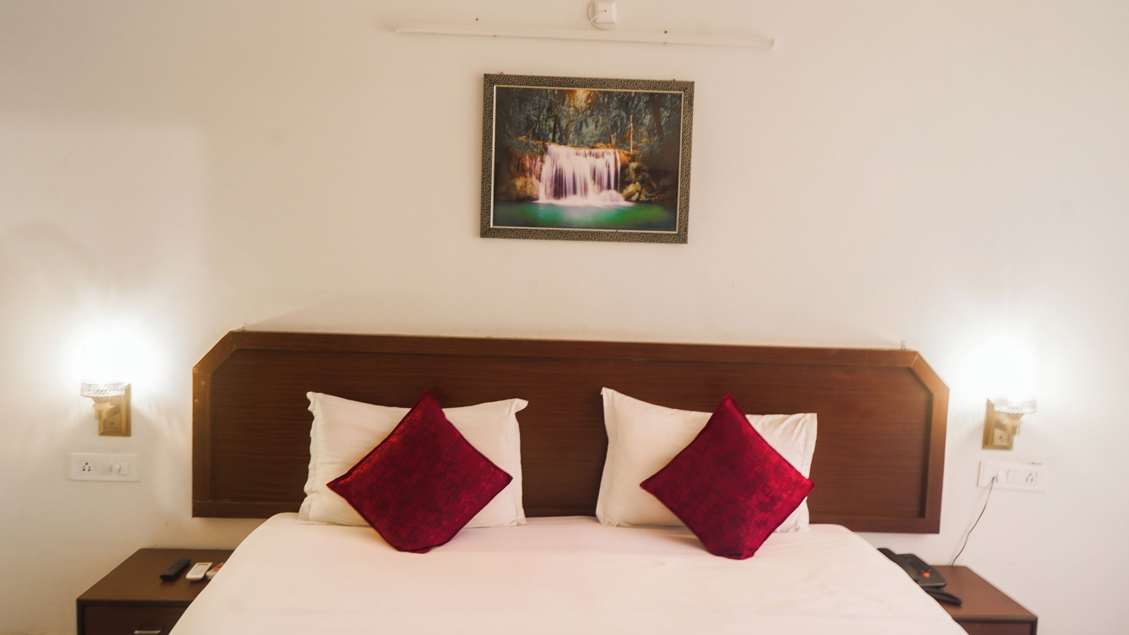 Una cama bien tendida con almohadas blancas y rojas, un cabecero de madera y un cuadro enmarcado de una cascada en la pared.