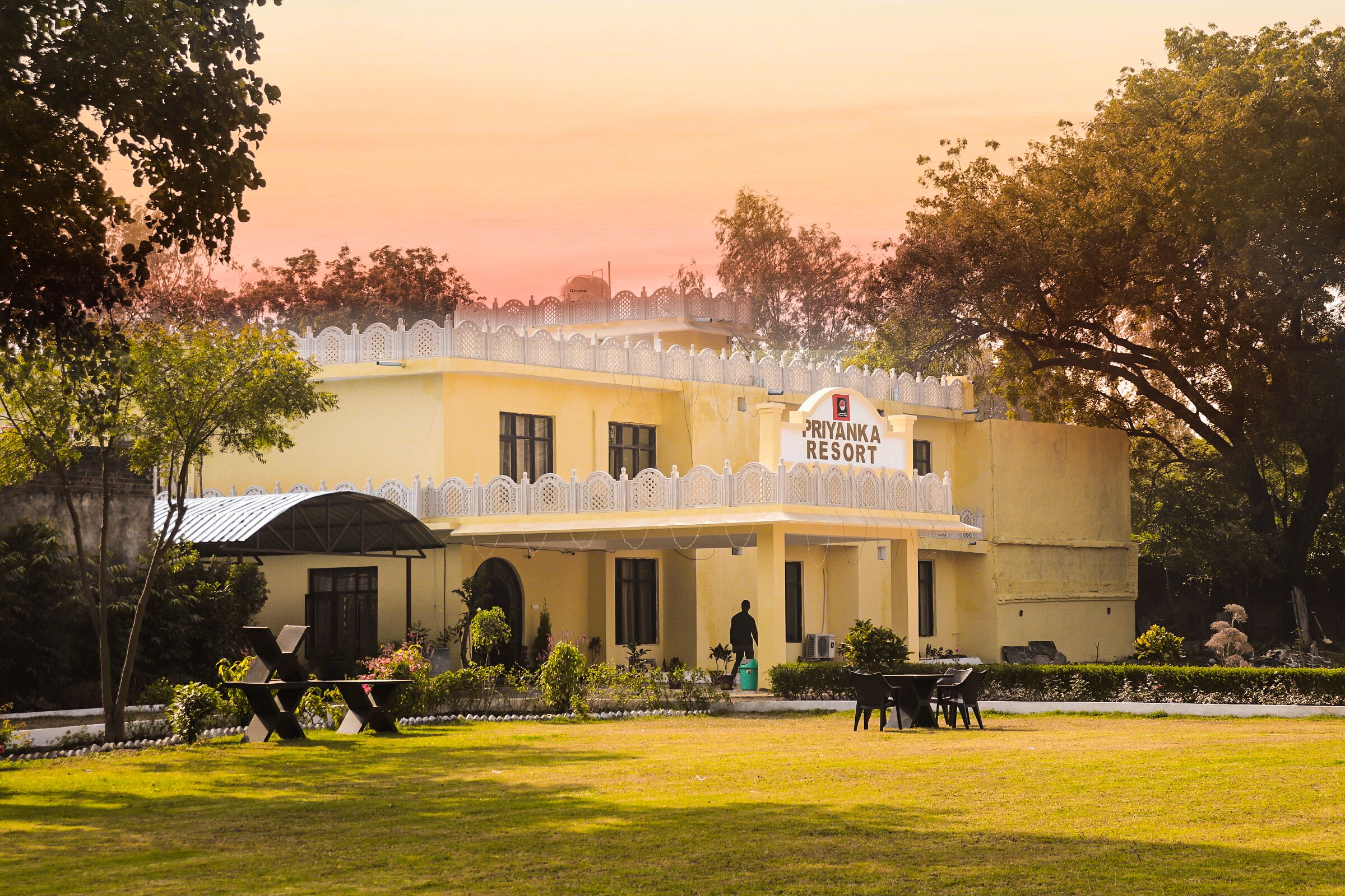 A building labeled "Priyanka Resort" with a covered entrance, a lawn, and outdoor seating.
