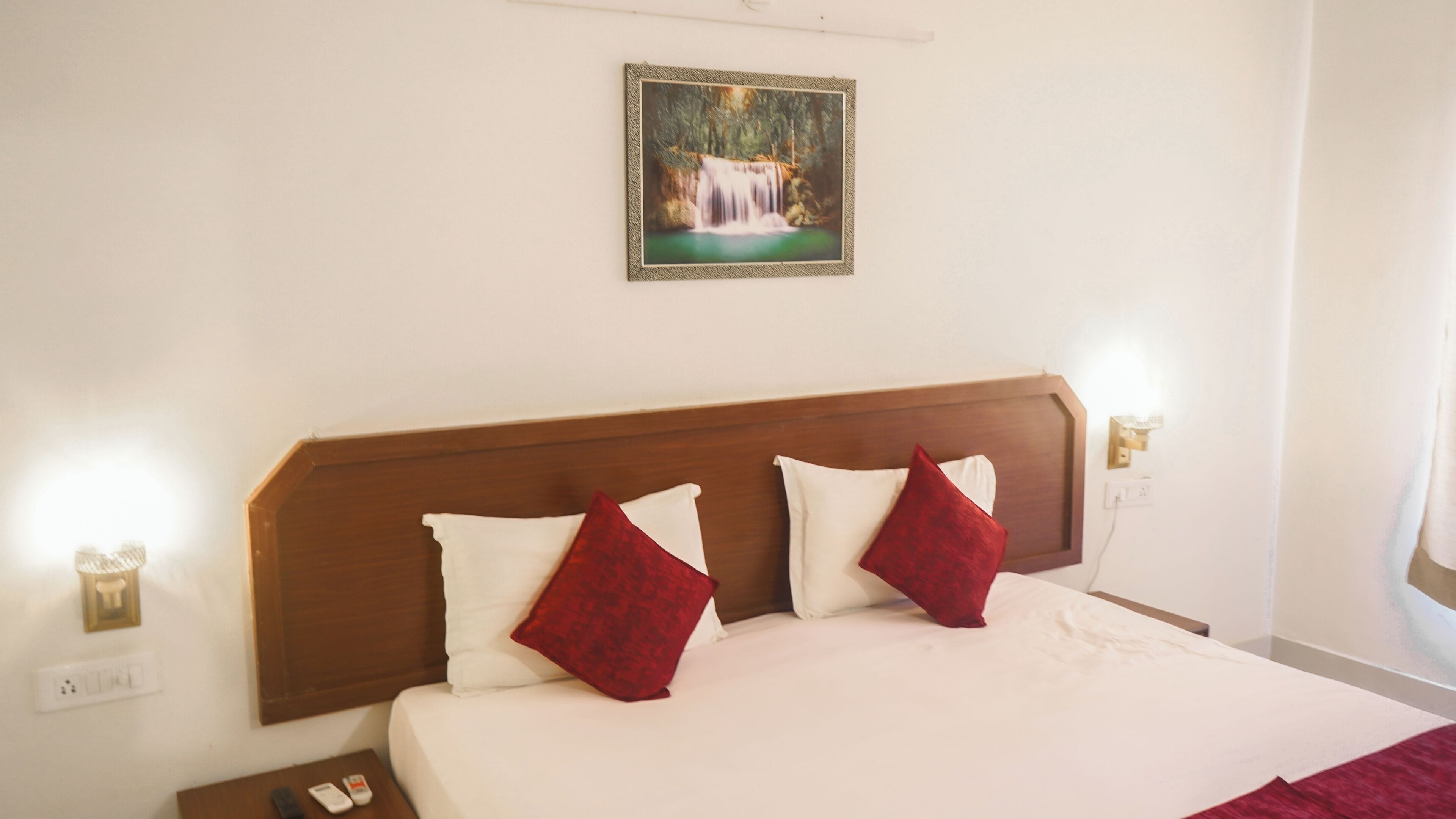 Habitación de hotel con cabecera de madera, ropa de cama blanca y almohadas rojas. Hay un cuadro de una cascada en la pared.