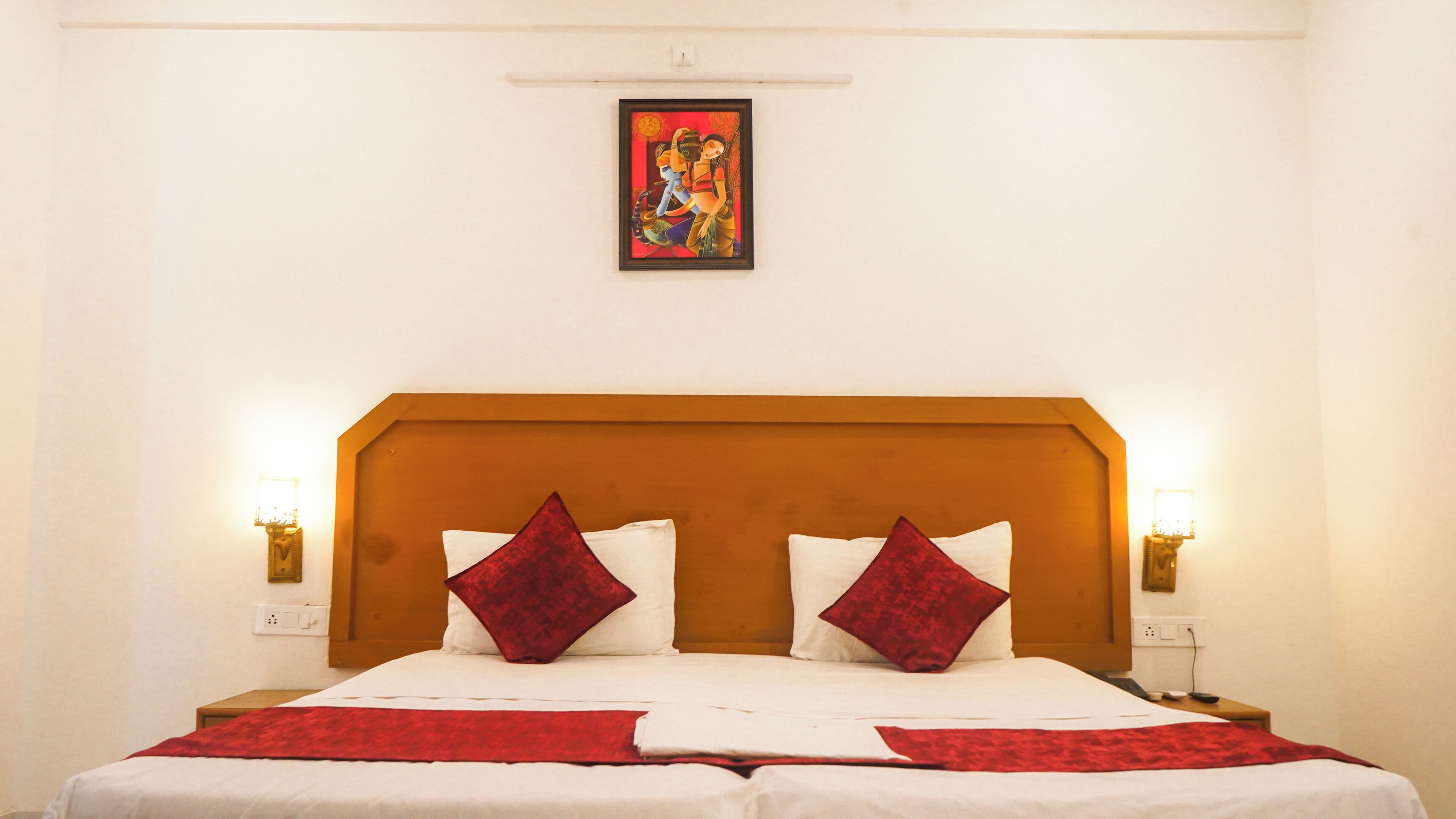 A hotel room with a wooden headboard, two red pillows, a white bedspread, and a painting above the bed.