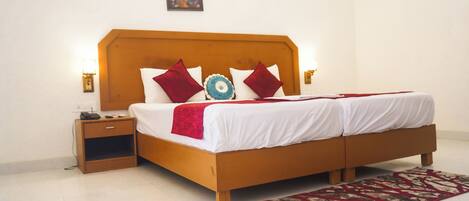 Cama doble con cabecera de madera, ropa de cama blanca y almohadas azules y rojas. Hay una mesita de noche con un teléfono y una lámpara a cada lado de la cama. Una alfombra estampada está en el suelo.