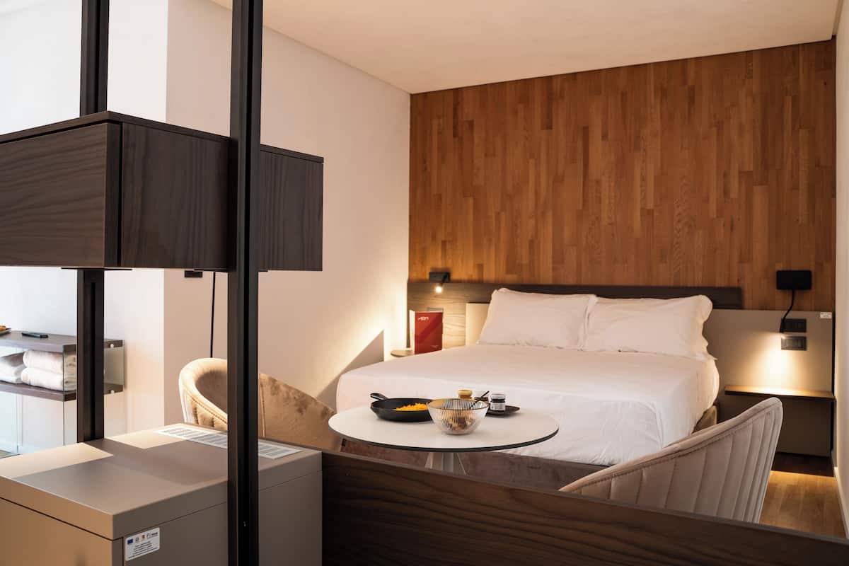 A hotel room with a bed, bedside table, and a small round table with a bowl and spoon.