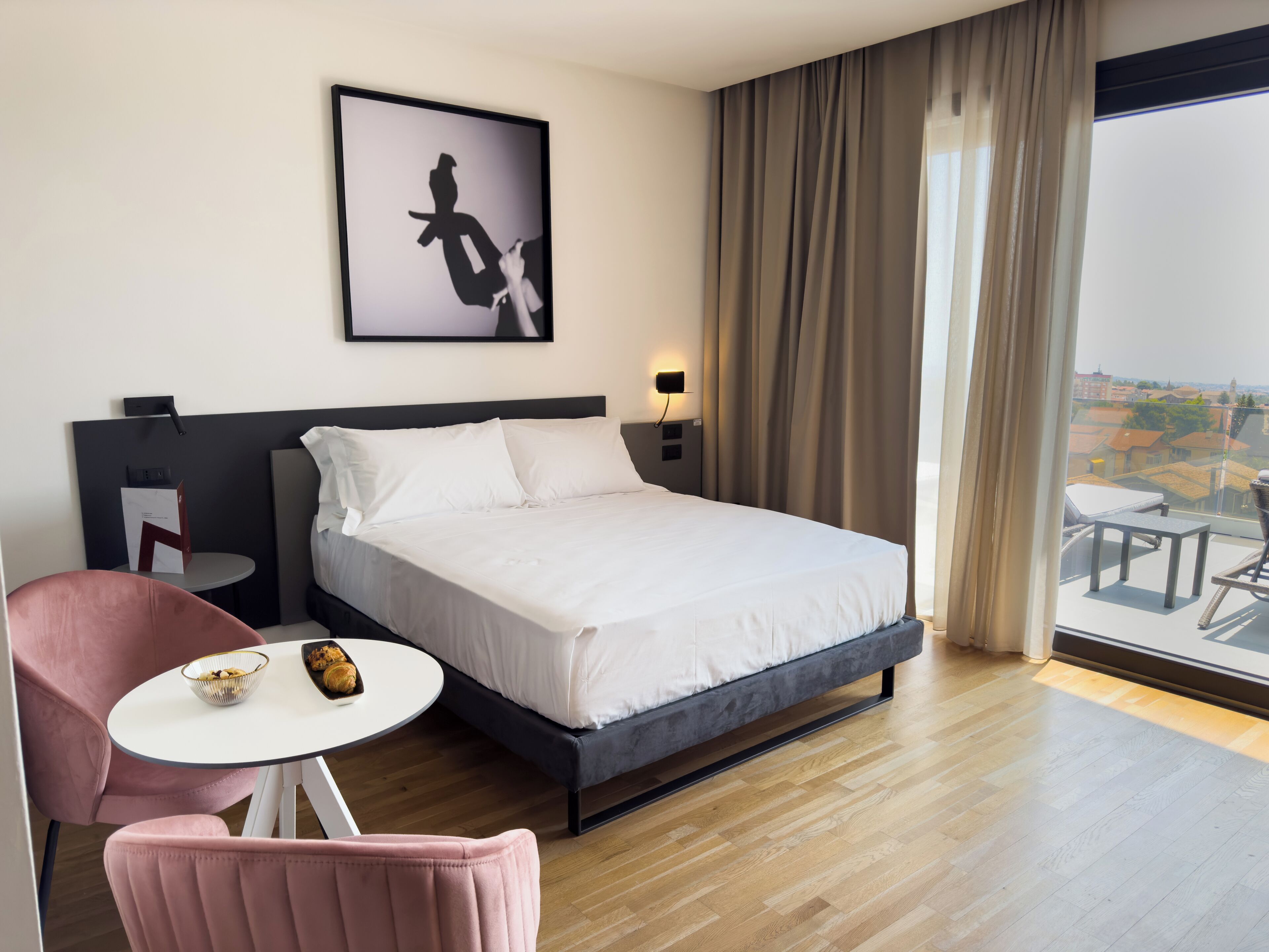 A hotel room with a bed, a small table with a bowl of pastries, a pink chair, a framed artwork on the wall, and a balcony with a view.