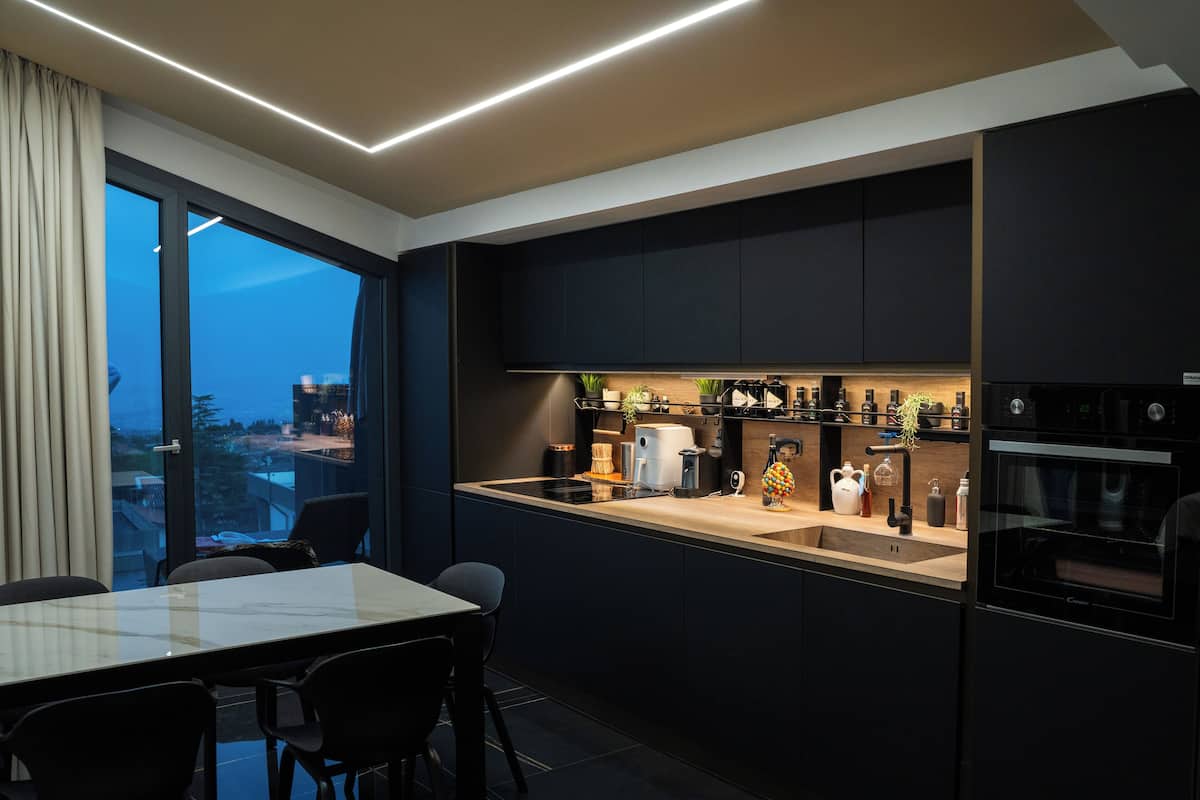 A modern kitchen with a large window, dark cabinetry, and a dining area with a table and chairs.
