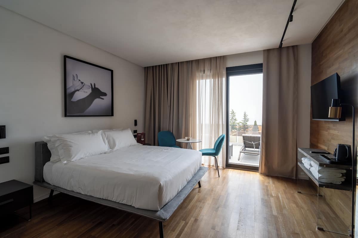 A modern hotel room with a large bed, a desk with a chair, a small table, and a view of the outdoors through a sliding glass door.