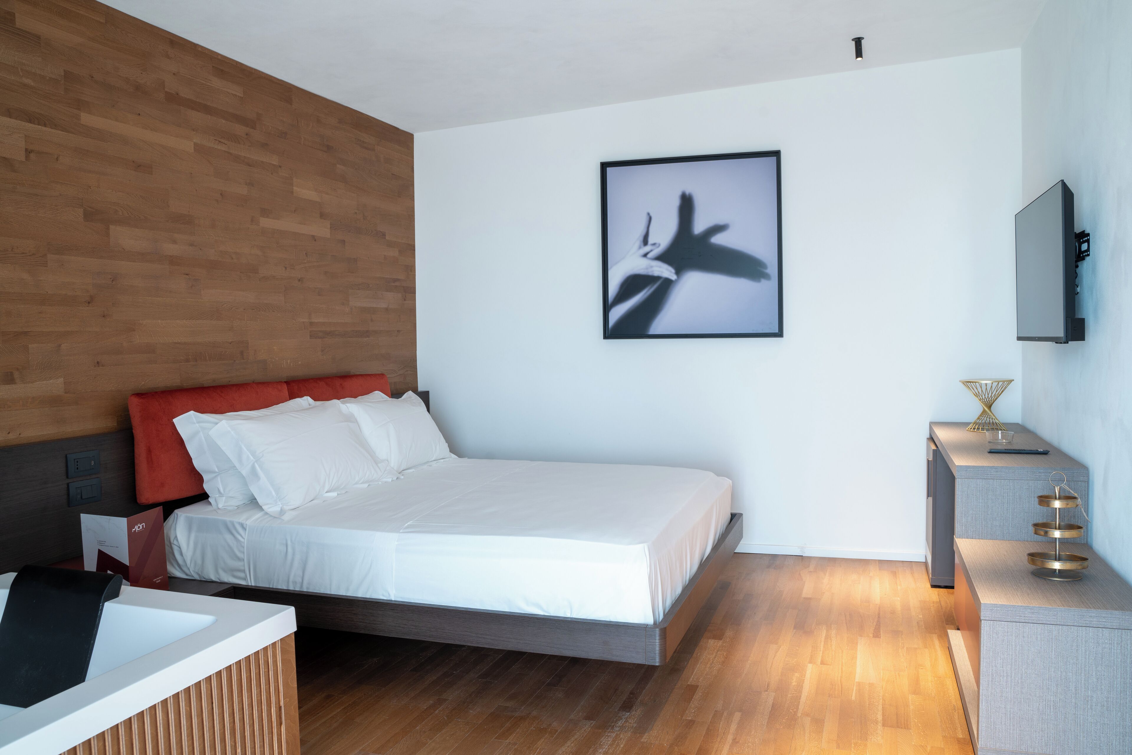 A modern hotel room with a wooden wall, a bed with white bedding, a mounted TV, and a bedside table with a tray.