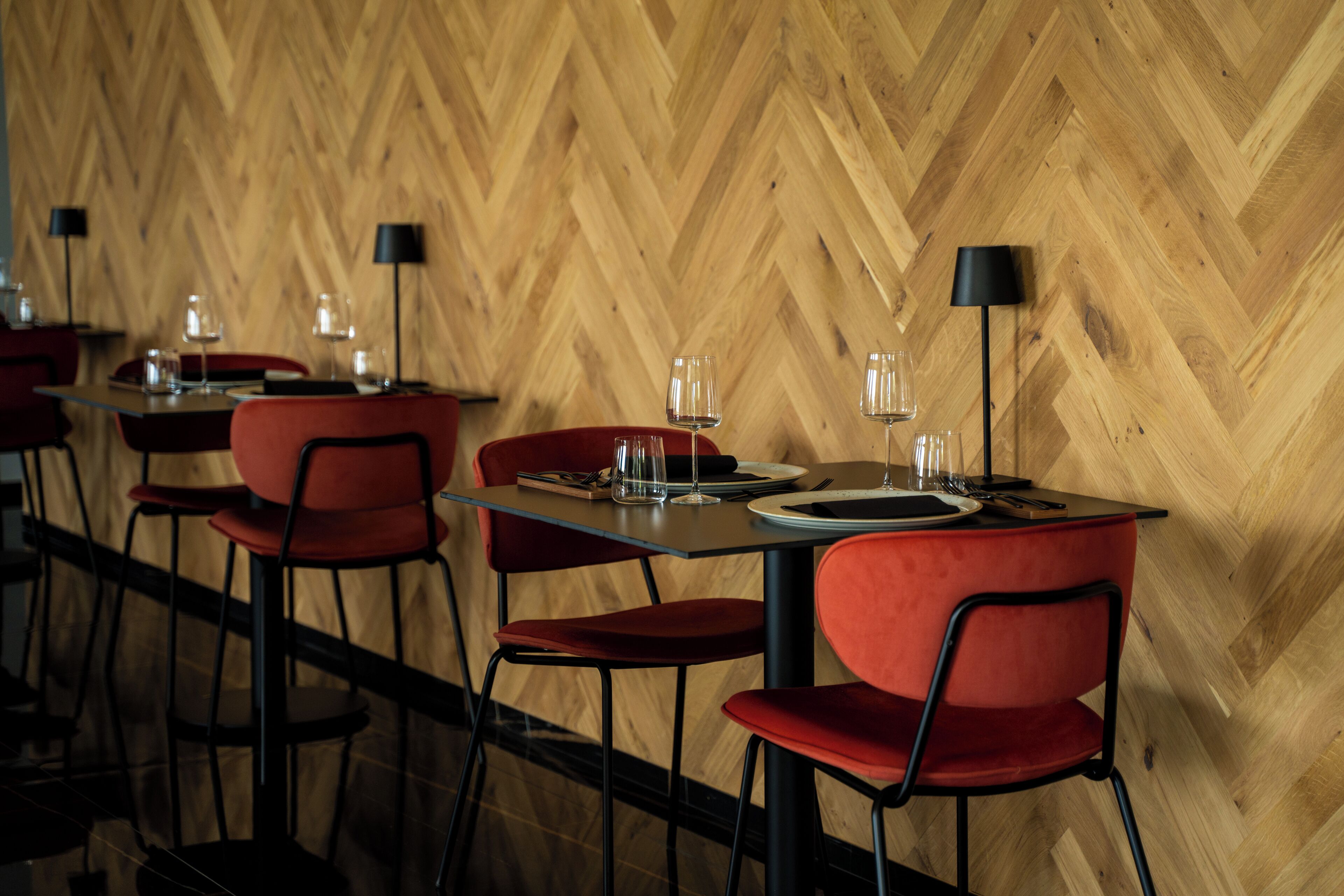 A restaurant with wooden herringbone flooring, red upholstered chairs, and black table lamps.