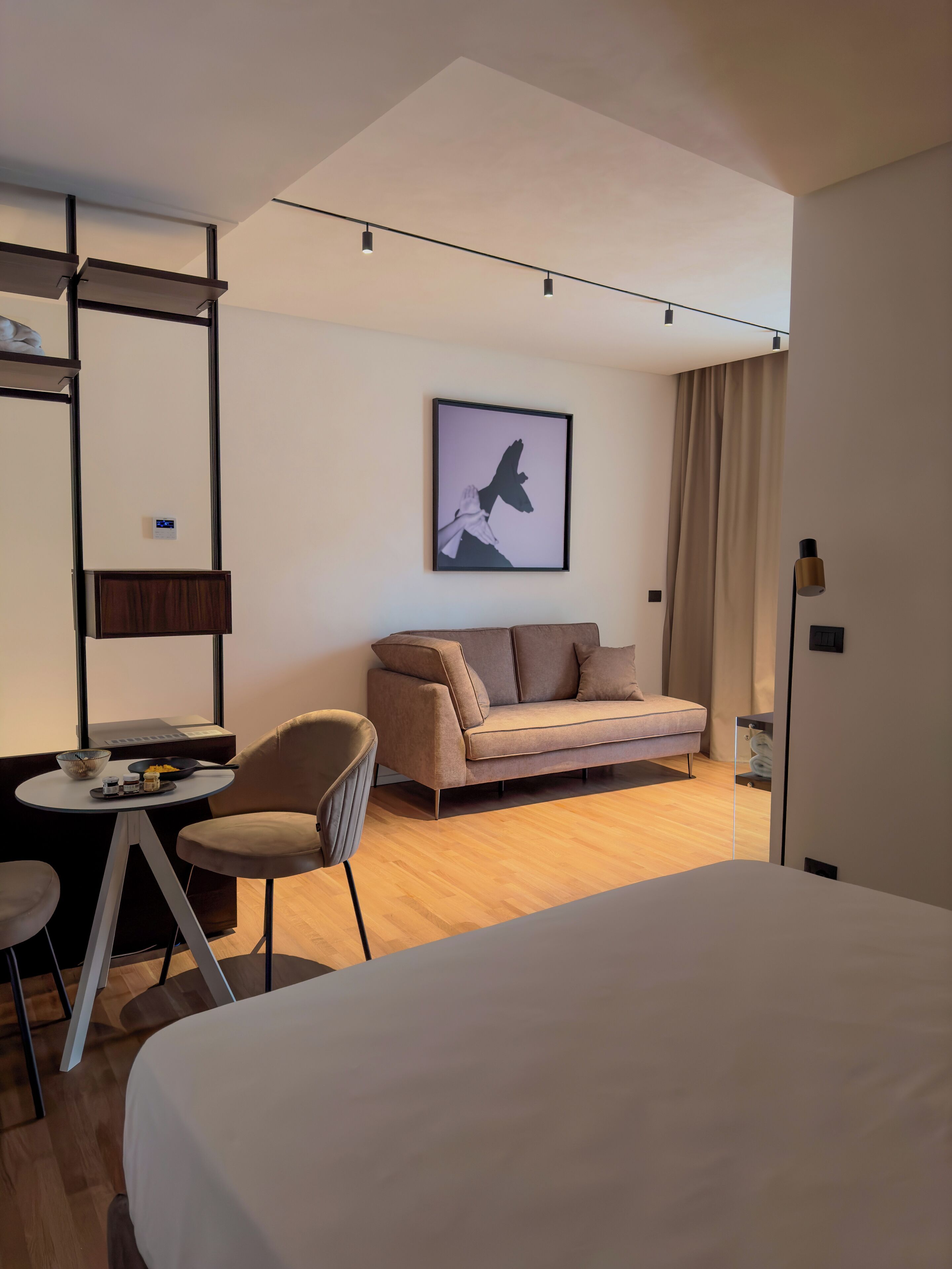 Une chambre d’hôtel moderne comprenant un lit, un canapé, une chaise, une petite table avec un bol et des condiments, un tableau encadré au mur et une fenêtre avec des rideaux.