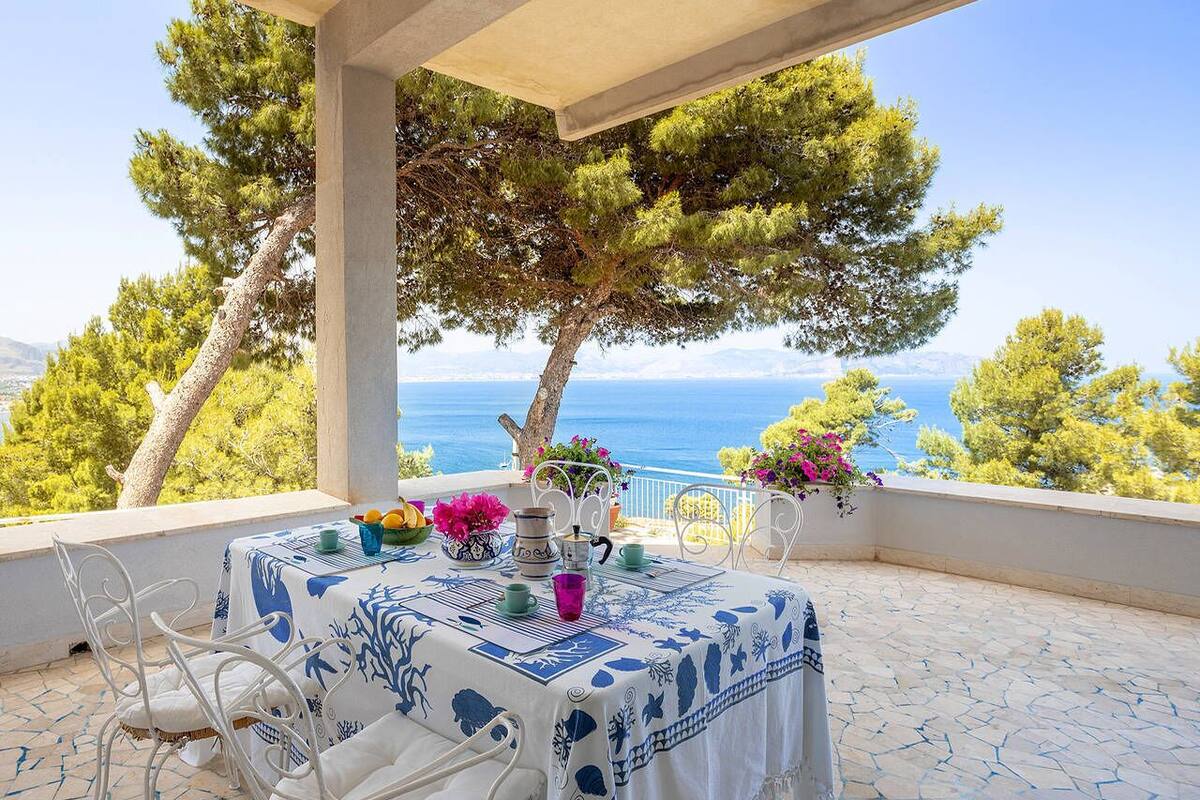 A terrace with a table set for dining, overlooking a scenic view of the sea and trees.
