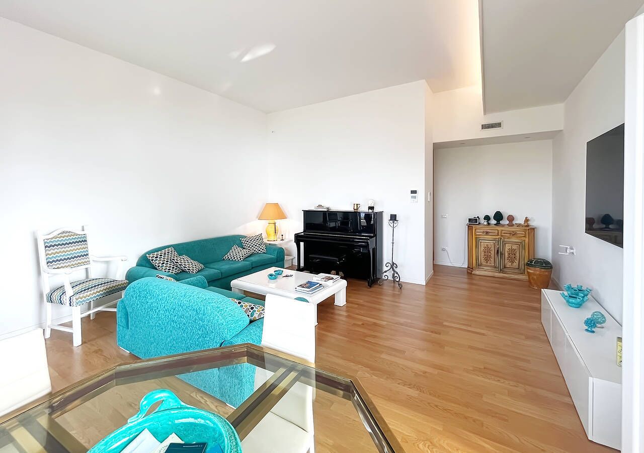 A living room with a teal sofa, a white armchair, a glass coffee table, a black piano, and a wooden cabinet.