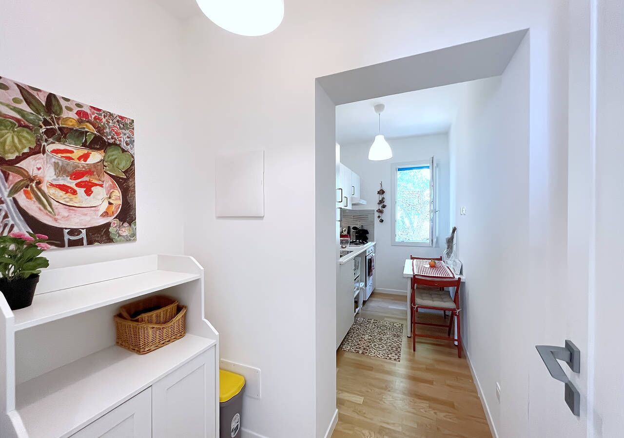 Un corridoio stretto che conduce a una cucina con mobili bianchi, una zona pranzo con una sedia rossa e un quadro appeso al muro.