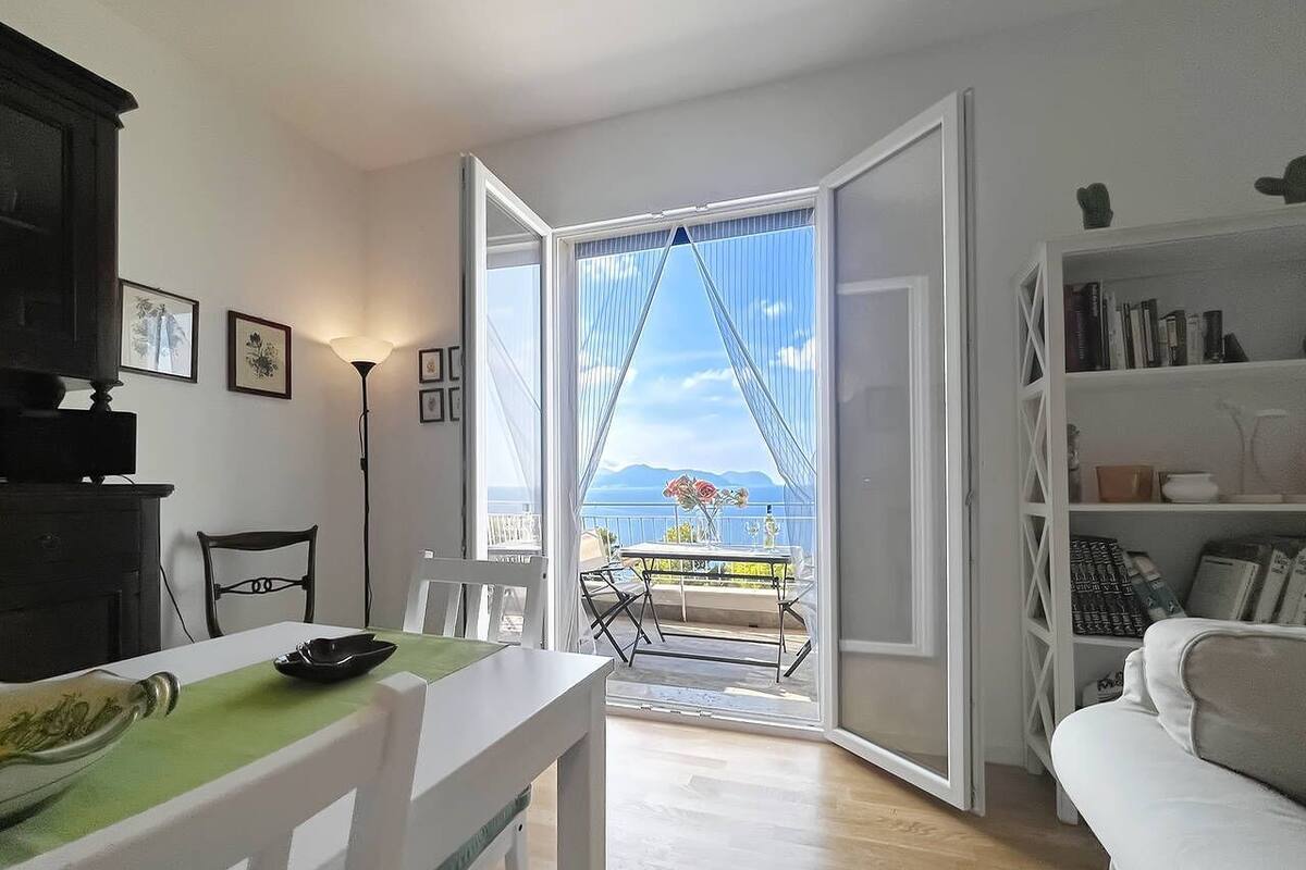 A modern living room with a dining area, a white sofa, a bookshelf, and a view of the sea through an open door.