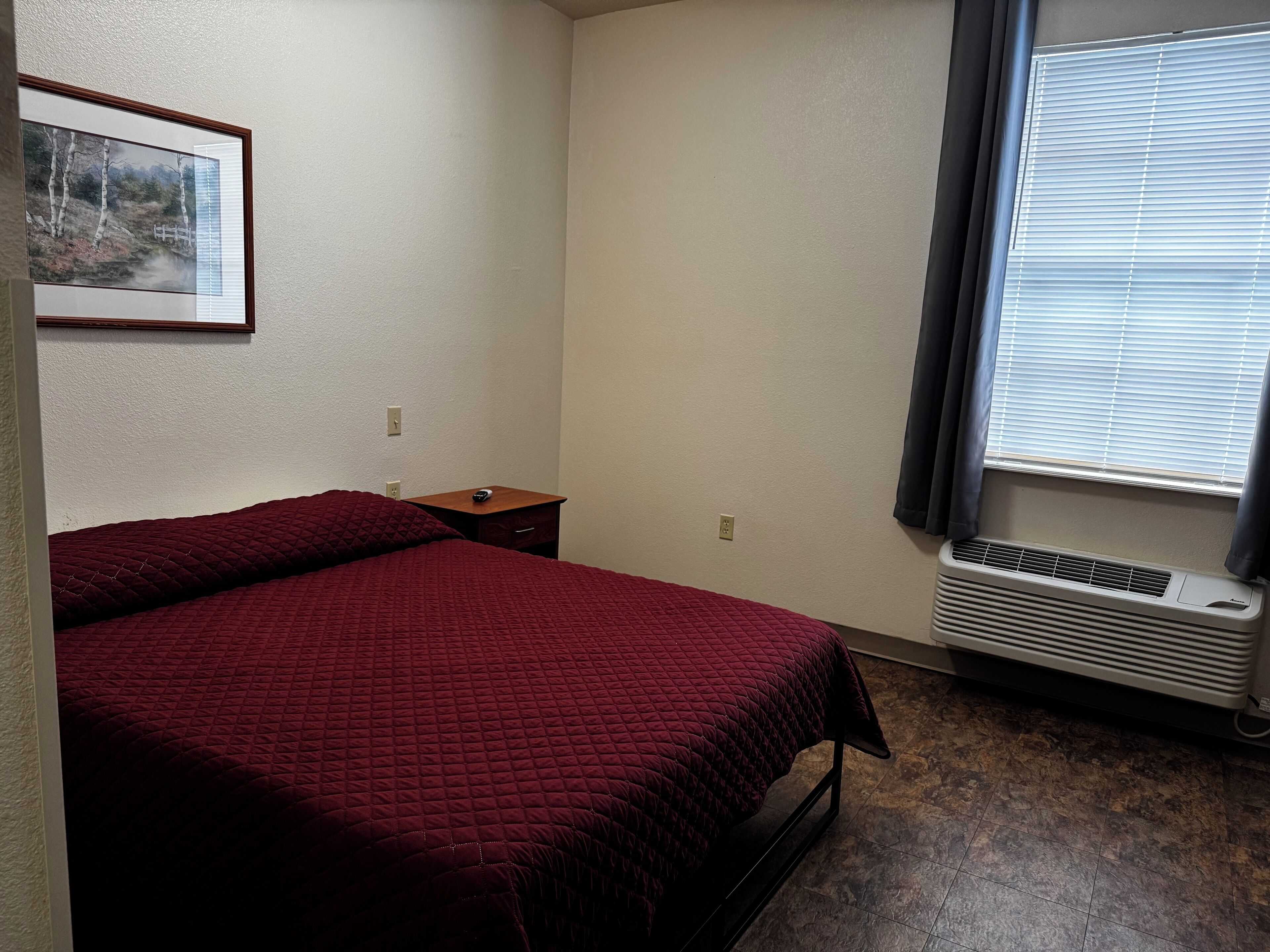 A small, neatly arranged bedroom with a single bed, a nightstand, a framed picture on the wall, and a window with blinds.