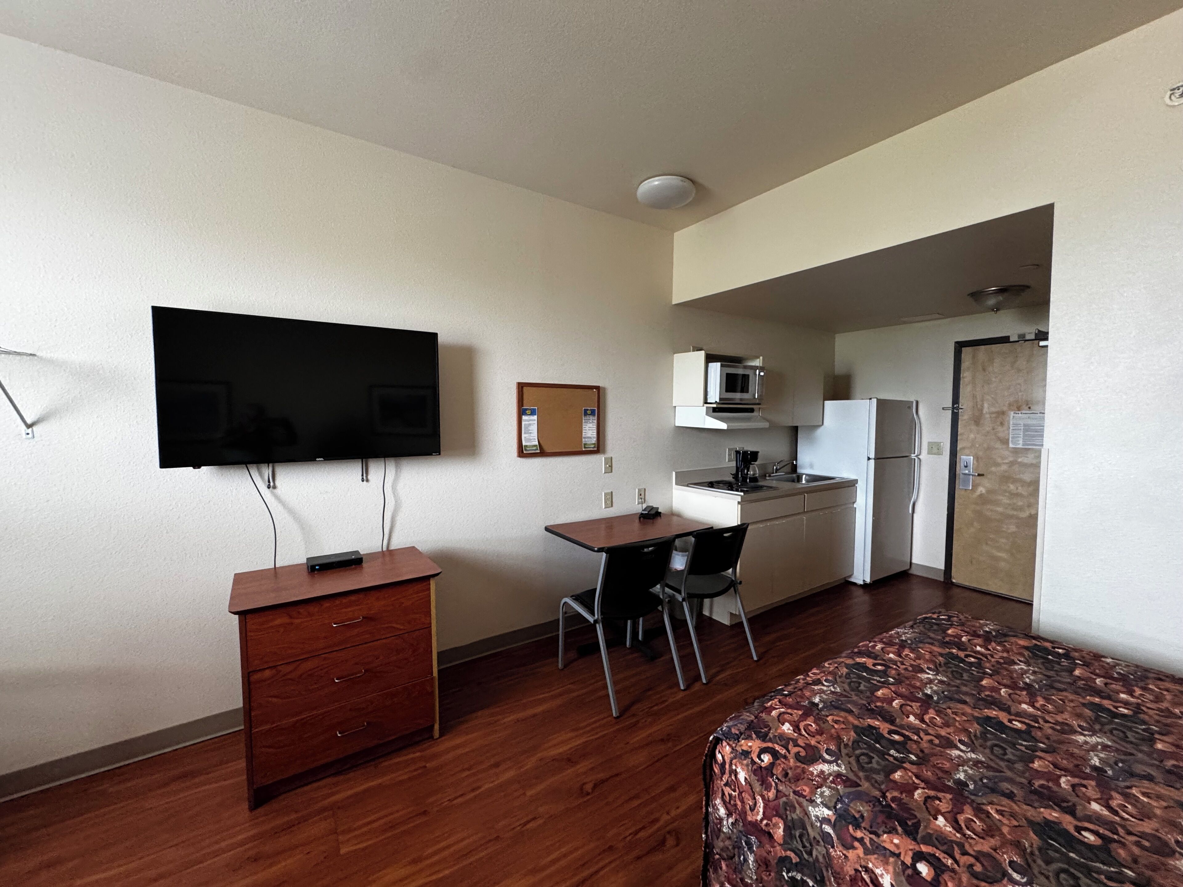 A compact hotel room with a wooden desk, a flat-screen TV, a small kitchenette, and a bed with a patterned cover.