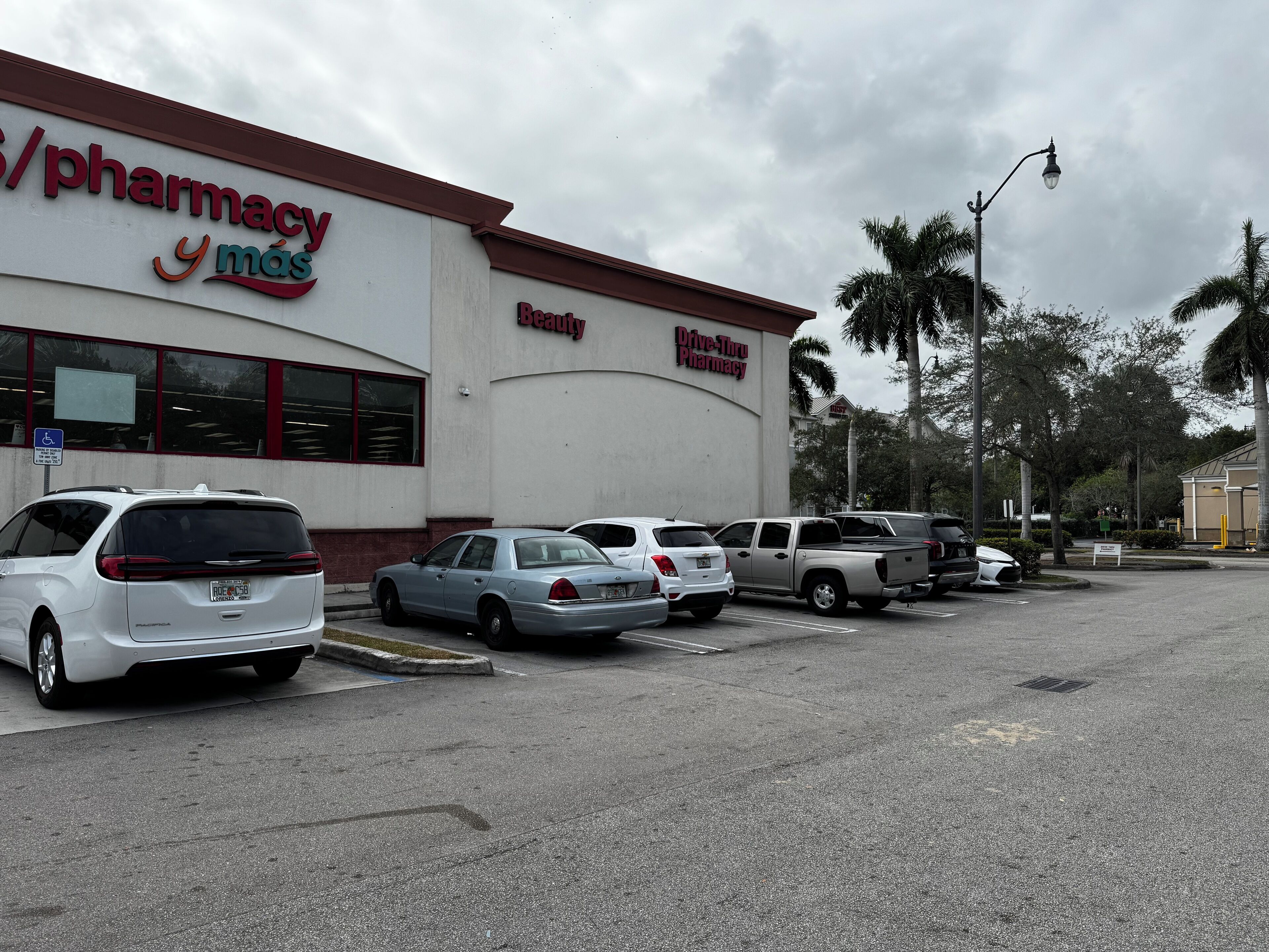 A pharmacy with a drive-thru service, parked cars, and palm trees in the background.