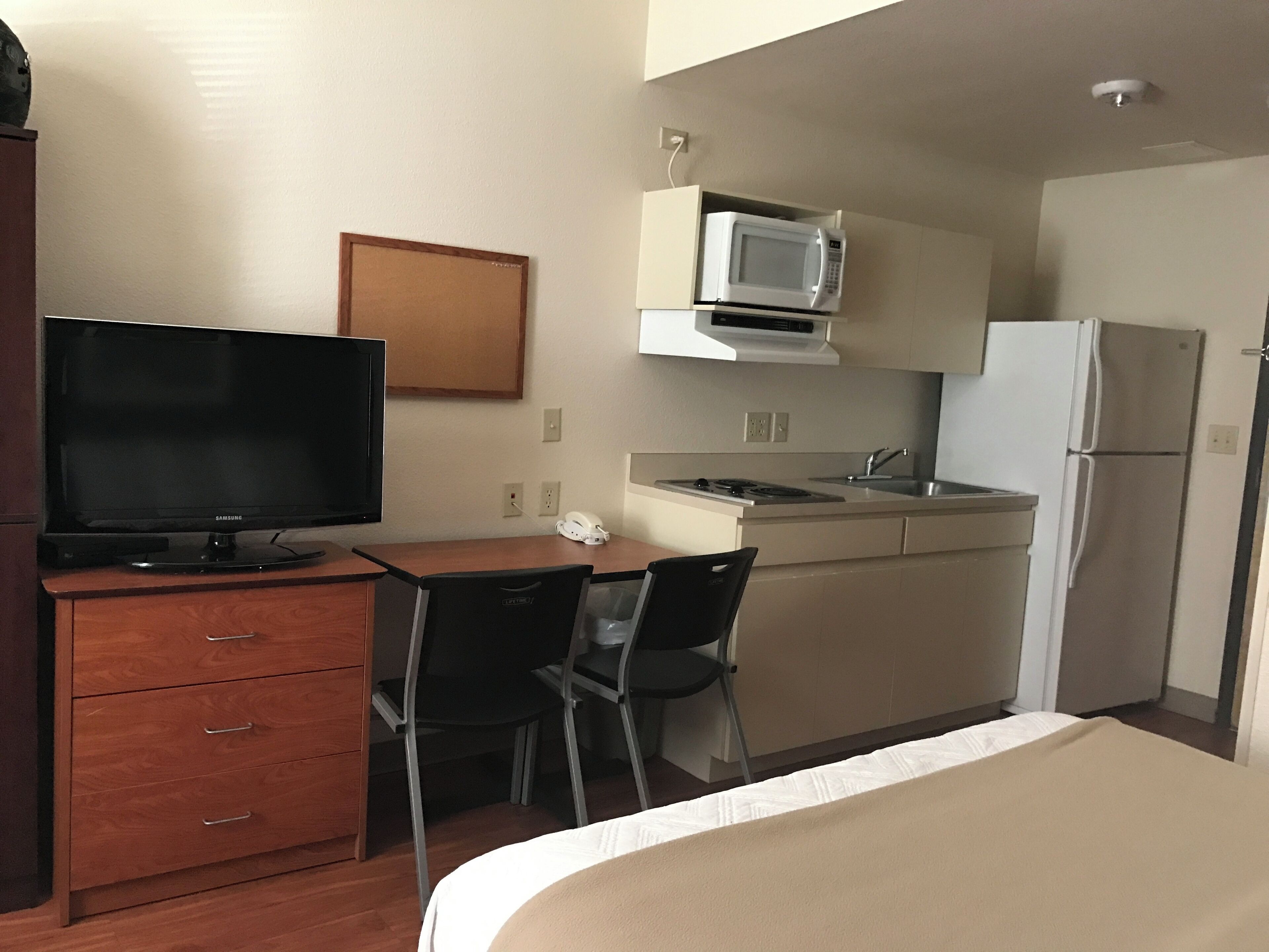 A compact hotel room with a bed, a desk with a television, a kitchenette with a microwave and refrigerator, and a chair.