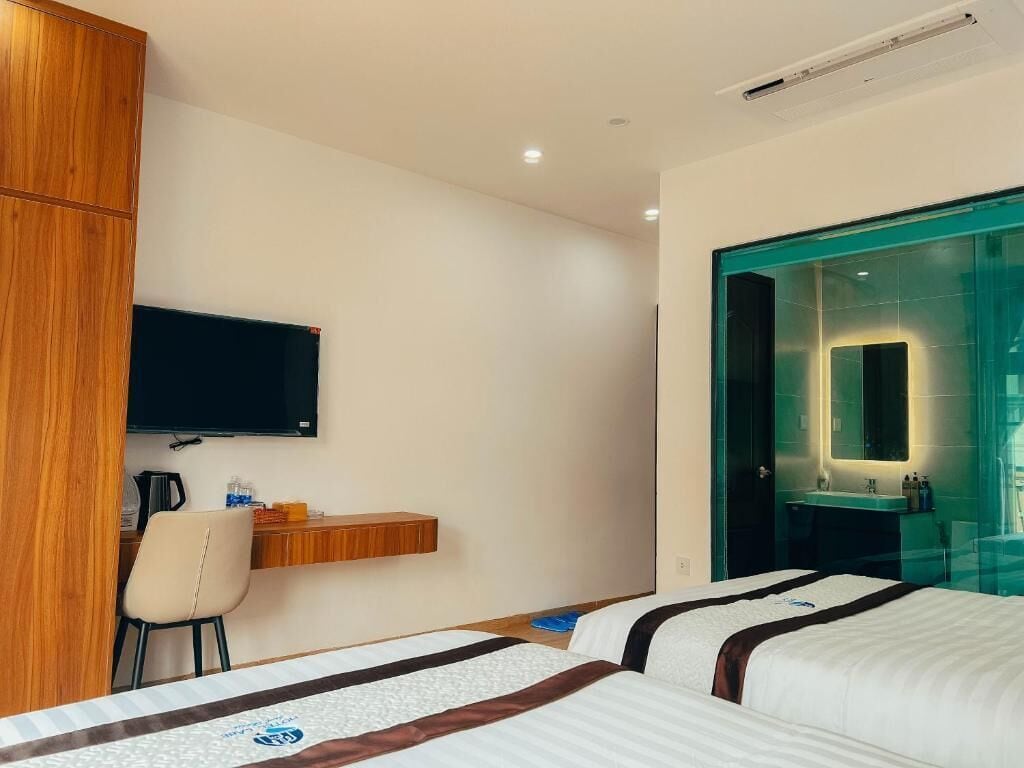 Ein Hotelzimmer mit zwei Betten, einem an der Wand befestigten Fernseher, einem Schreibtisch mit Stuhl und einem Badezimmer mit Glastür.