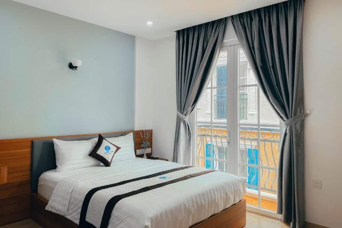 Une chambre d’hôtel avec un lit, une table de chevet, une lampe fixée au mur et une vue sur un balcon doté d’une balustrade.