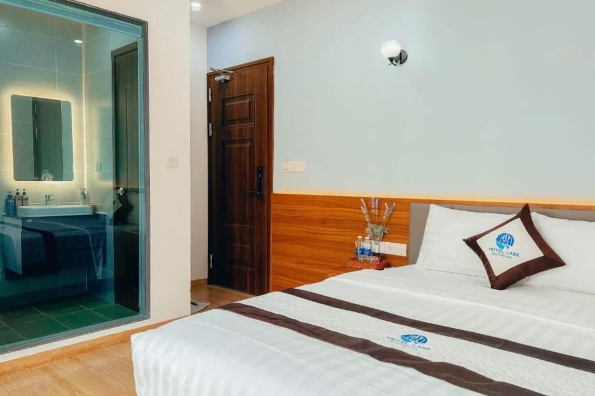 Une chambre d’hôtel avec un lit, une tête de lit en bois, une table de chevet avec une lampe et une cabine de douche en verre.