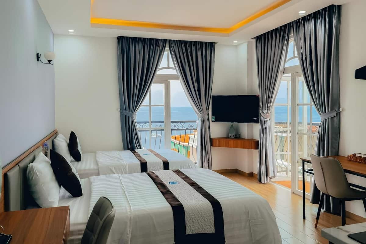 Une chambre d’hôtel avec deux lits, une télévision, un balcon avec vue sur la mer et un coin repas.