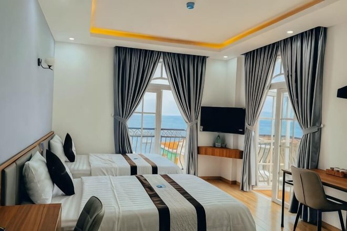 A hotel room with two beds, a desk, a chair, a TV, and a balcony with a view.
