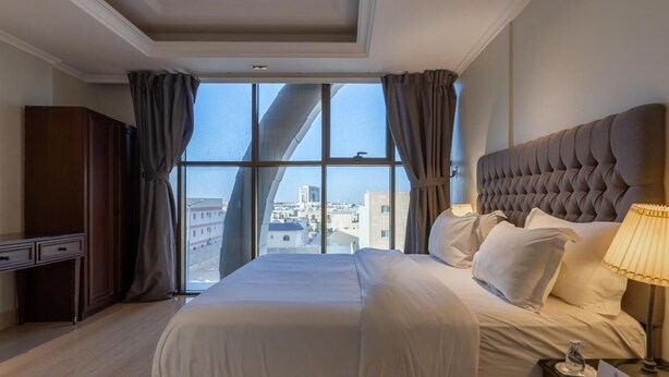 A hotel room with a large bed, a nightstand, a desk, and a view of the city through large windows. - Jazalah al falah serviced apartments  (Riyadh)
