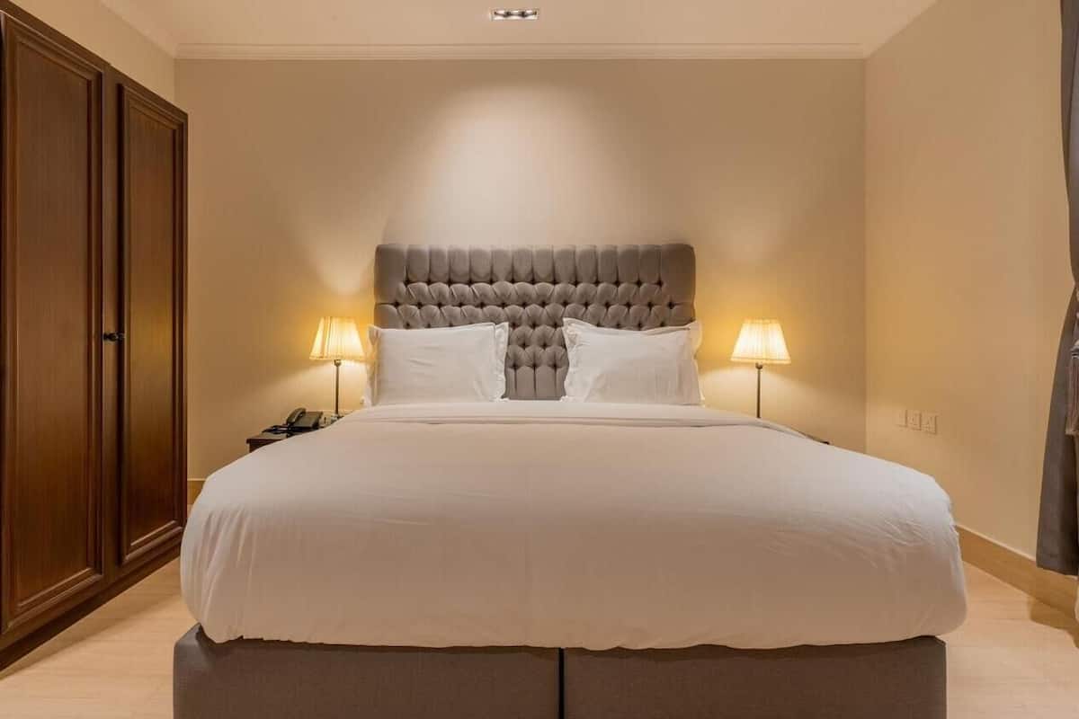 A large bed with a tufted headboard, two bedside lamps, and a wooden wardrobe.