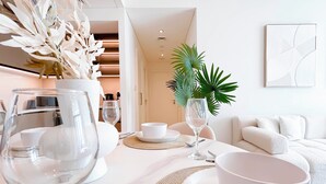 A modern, well-lit dining area with a white table set for two, featuring a clear glass wine glass, a white bowl, and a potted plant.