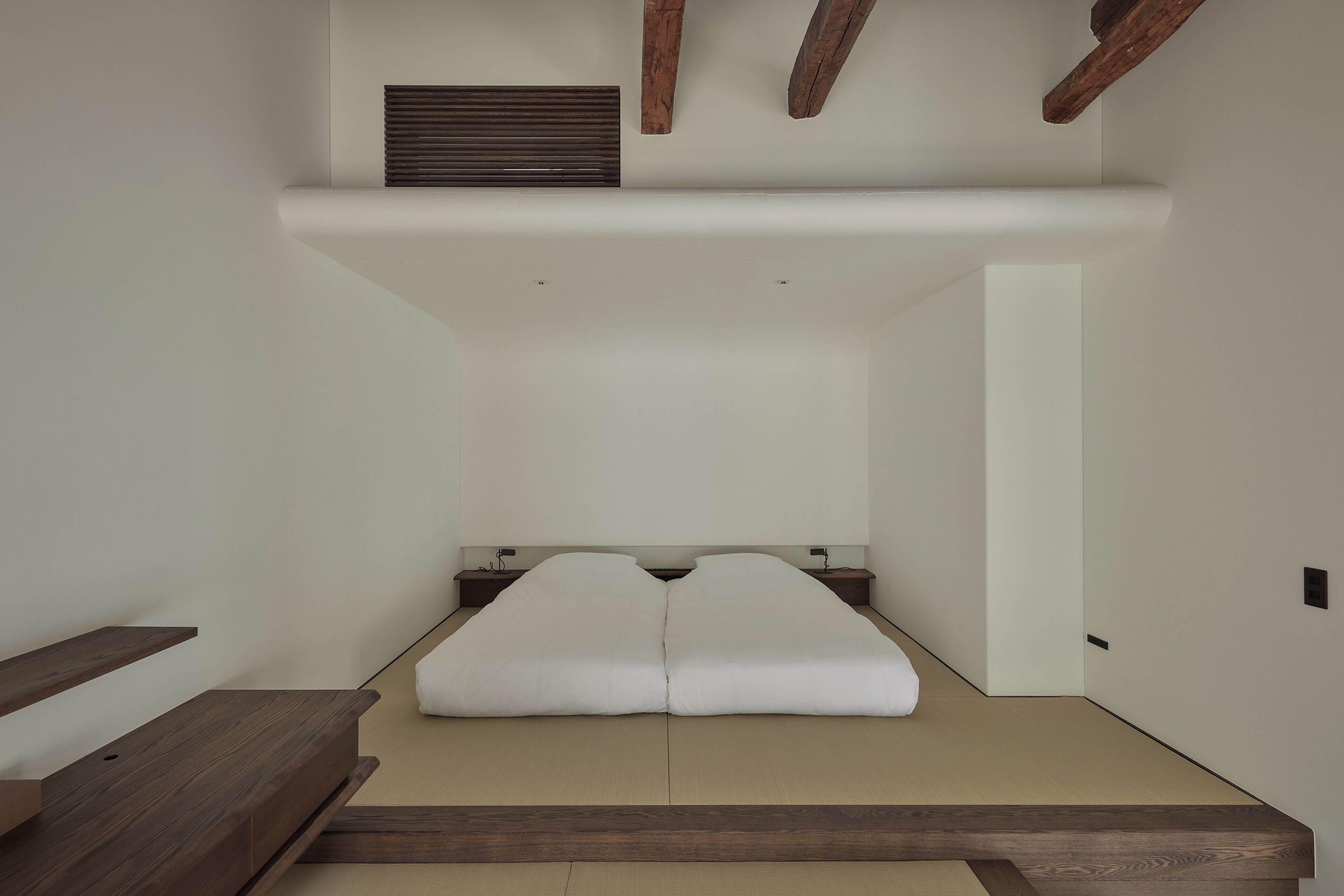 Habitación minimalista con una cama baja sobre plataforma, vigas de madera y una mesita de noche pequeña.