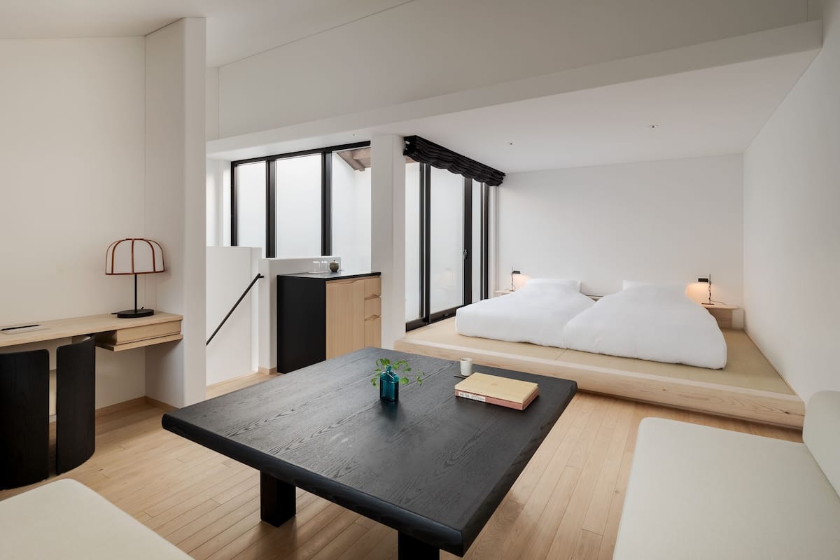 Et moderne, minimalistisk rum med trægulv, et sort bord med en plante, en hvid sofa med puder og et skrivebord med en lampe.