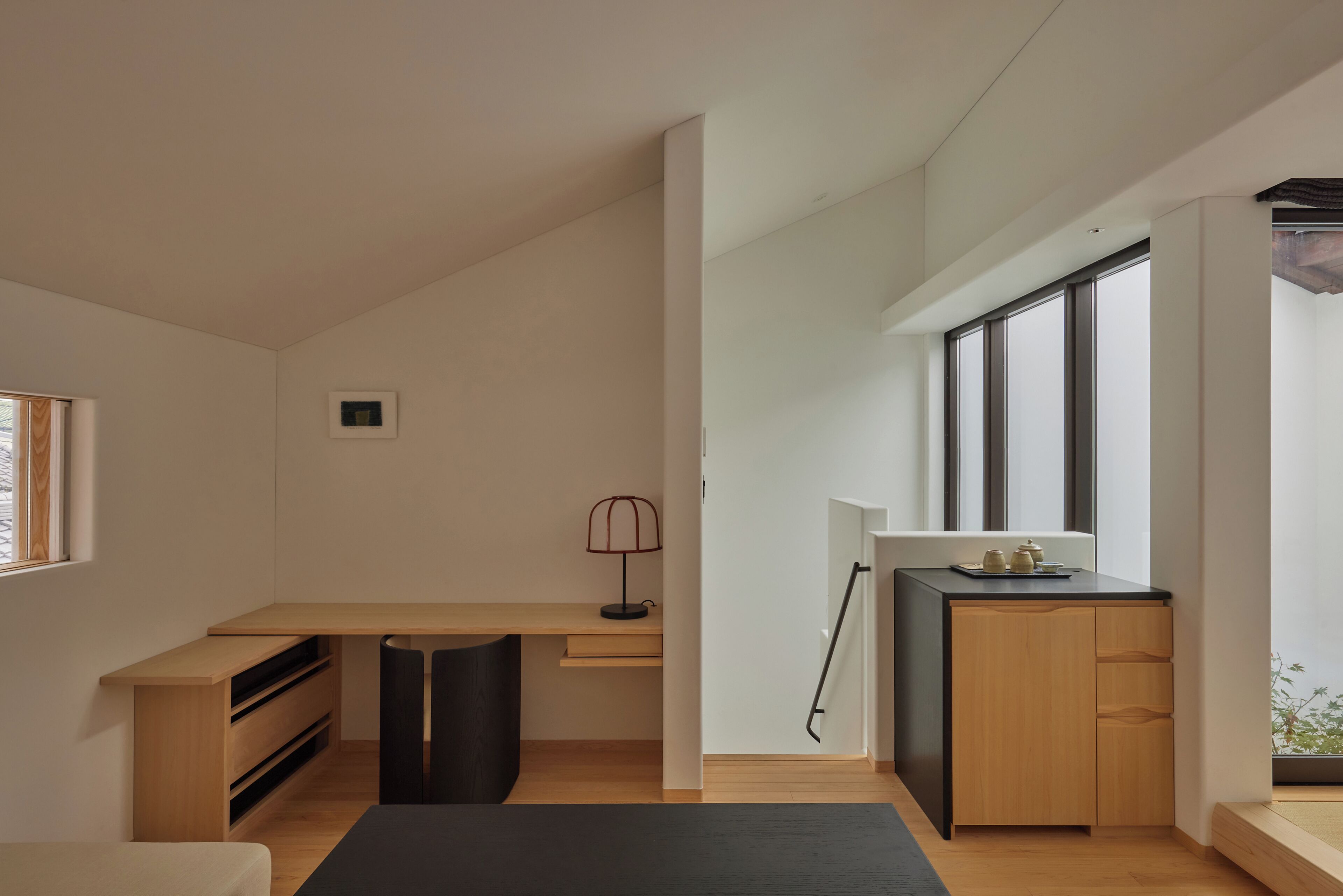 A modern, minimalist room with a wooden desk, a black chair, a small lamp, and a view through large windows.